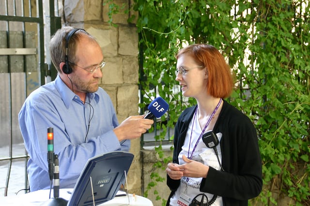 Muž s mikrofonem vede rozhovor se ženou; oba stojí proti sobě a soustředí se na výměnu.